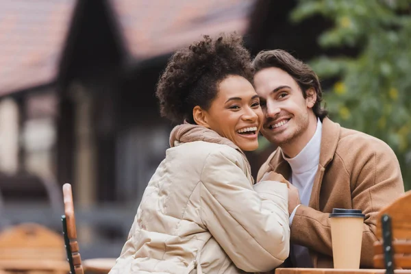 Heureux couple interracial en manteaux tenant la main près de tasse de papier sur la table — Photo de stock