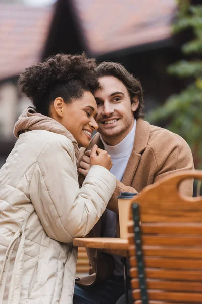 Joyful interracial couple in coats holding hands near paper cup on table — Stock Photo