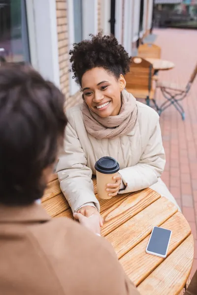 Felice donna afroamericana che si tiene per mano con ragazzo sfocato vicino smartphone e tazza di carta — Foto stock