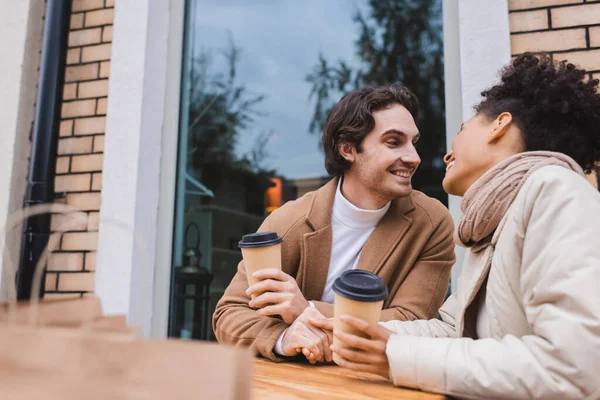 Joyeux couple interracial en manteaux tenant les mains et les tasses en papier près des sacs à provisions flous — Photo de stock