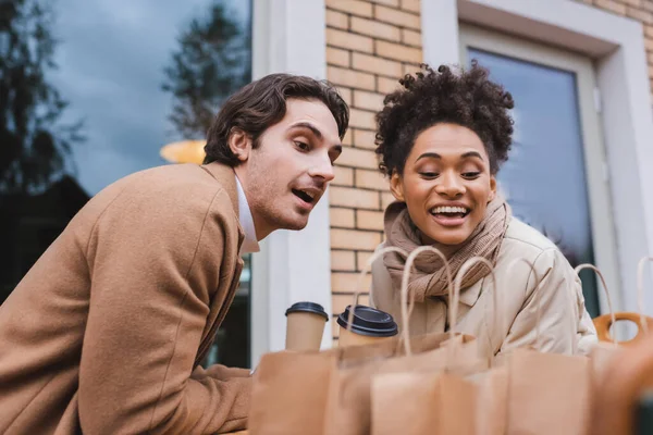 Alegre pareja interracial en abrigos sosteniendo vasos de papel y mirando borrosas bolsas de compras - foto de stock