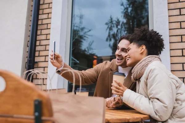 Happy interracial couple taking selfie near paper cups and blurred shopping bags — Stock Photo