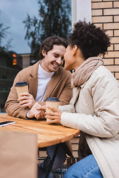 Glückliches gemischtrassiges Paar Händchen haltend in der Nähe von Pappbechern und verschwommener Einkaufstasche — Stockfoto