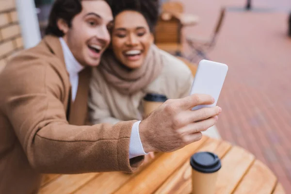 Borrosa y feliz interracial pareja tomando selfie cerca de papel tazas - foto de stock