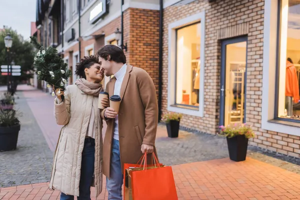 Feliz casal interracial com café para ir, sacos de compras de Natal e pequeno pinho perto do shopping — Fotografia de Stock