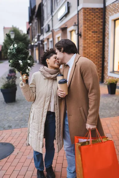 Allegra coppia interrazziale con caffè da andare, borse della spesa di Natale e piccolo pino vicino al centro commerciale — Foto stock