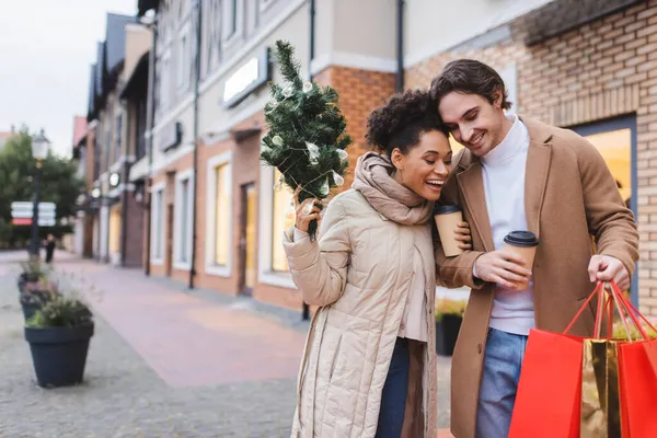 Stupito coppia interrazziale con caffè da andare, borse della spesa di Natale e piccolo pino vicino al centro commerciale — Foto stock