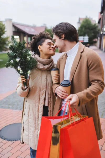 Smiling interracial couple with coffee to go, christmas shopping bags and small pine outdoors — Stock Photo