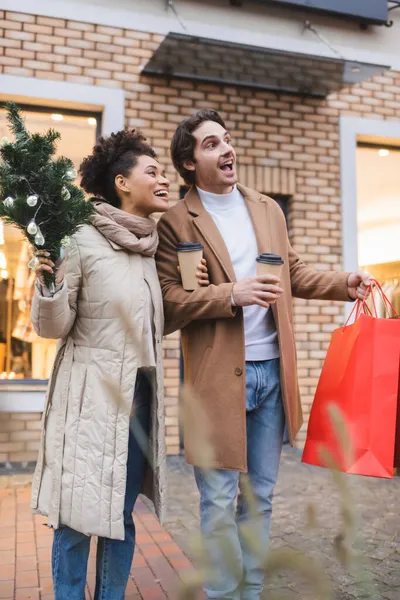 Animado casal multiétnico com café para ir, saco de compras de Natal e pequeno pinheiro perto do shopping — Fotografia de Stock