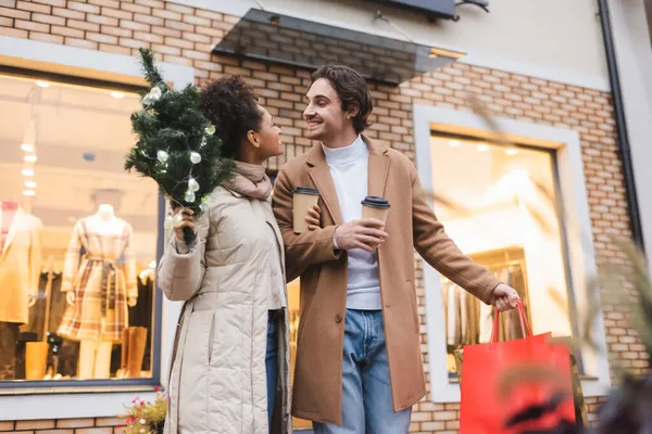 Felice coppia multietnica con caffè da asporto, borse della spesa natalizie e piccolo abete vicino al centro commerciale — Foto stock