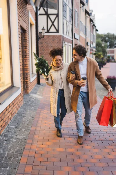 Feliz casal multiétnico com café para ir, sacos de compras de Natal e pinho andando perto do shopping — Fotografia de Stock