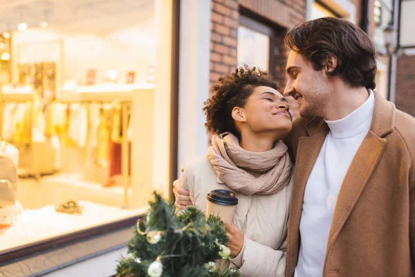 Feliz pareja multiétnica con los ojos cerrados sosteniendo café para ir y pino cerca del centro comercial — Stock Photo