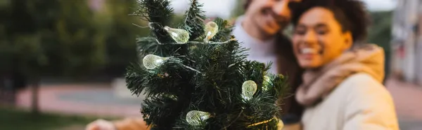 Coppia interrazziale offuscata vicino rami di pino di Natale con luci ghirlanda, banner — Foto stock