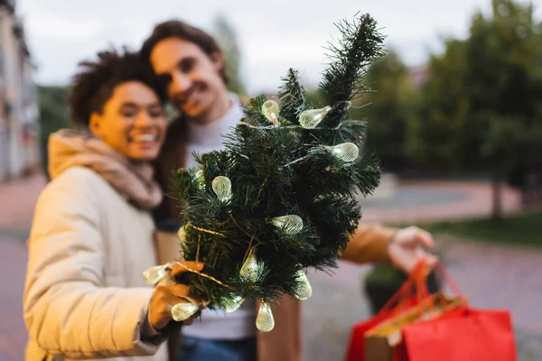 Femme afro-américaine floue tenant petit pin de Noël avec guirlande lumières près du petit ami — Photo de stock