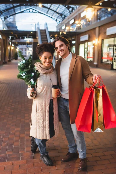 Heureux couple multiethnique tenant café à emporter, sacs de Noël et pin en soirée — Photo de stock