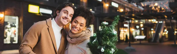 Happy multiethnic couple holding small christmas pine in evening, banner — Stock Photo