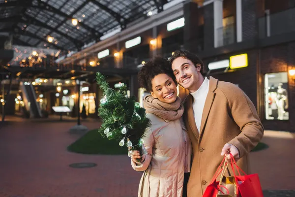 Feliz casal multiétnico segurando sacos de compras de Natal e pequeno pinho perto do shopping à noite — Fotografia de Stock