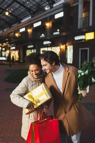 Felice coppia multietnica tenendo borse della spesa di Natale, confezione regalo e pino vicino al centro commerciale in serata — Foto stock