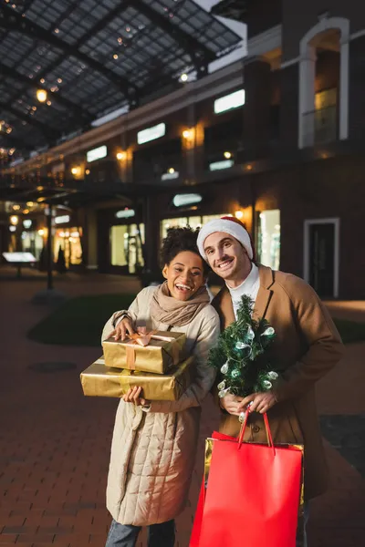 Hombre feliz en sombrero de santa celebración de bolsas de compras de Navidad y pino cerca de la novia afroamericana con regalos - foto de stock
