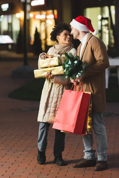 Fröhlicher Mann mit Weihnachtsmütze hält Weihnachts-Einkaufstüten und Kiefer in der Nähe der afrikanisch-amerikanischen Freundin mit Geschenken — Stockfoto