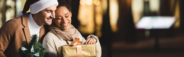 Happy man in santa hat holding christmas small pine near african american girlfriend with gift box, banner — Stock Photo