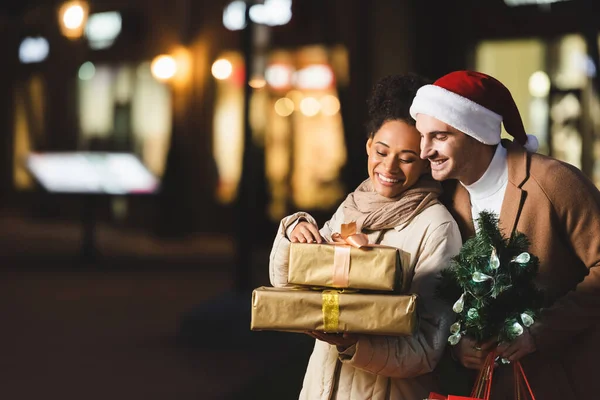 Счастливый человек в шляпе Санты, держащий рождественские сумки и маленькую сосну рядом с африканской американской подругой с подарочными коробками — стоковое фото