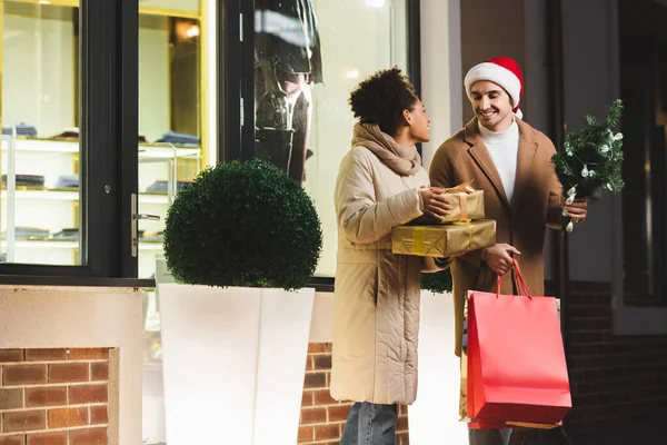 Glücklicher Mann in Weihnachtsmannmütze mit Einkaufstaschen und Kiefer in der Nähe einer afrikanisch-amerikanischen Freundin mit Geschenkboxen in der Nähe eines Einkaufszentrums — Stockfoto
