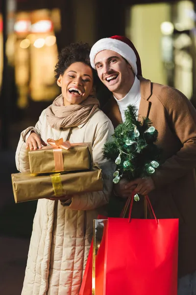 Hombre en sombrero de santa celebración de navidad bolsas de compras y pino cerca de novia afroamericana asombrada con cajas de regalo - foto de stock