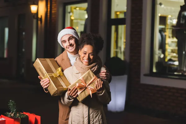 Happy man in santa hat and african american girlfriend holding christmas gift boxes — Stock Photo