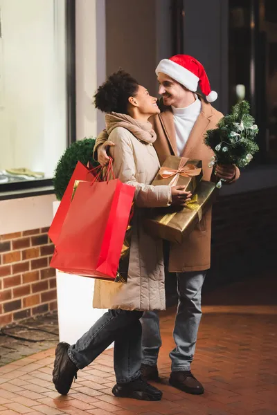 Homem feliz em santa chapéu segurando sacos de compras e abeto perto da namorada americana africana com caixas de presente — Fotografia de Stock