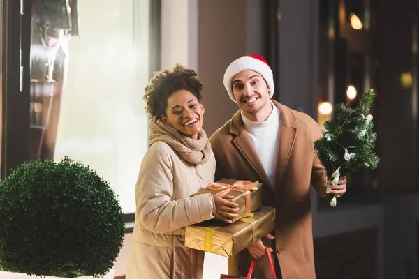 Hombre feliz en sombrero de santa celebración de bolsas de compras y abeto pequeño cerca de la novia afroamericana con cajas de regalo - foto de stock