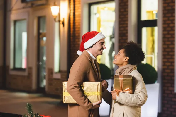 Hombre feliz en sombrero de santa y novia afroamericana mirándose mientras sostiene cajas de regalo de Navidad - foto de stock