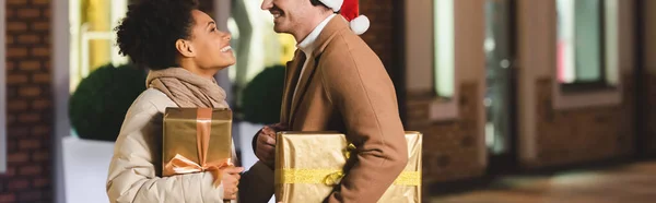 Щасливий чоловік в капелюсі Санти і афроамериканська дівчина дивиться один на одного, тримаючи різдвяні подарункові коробки, банер — стокове фото