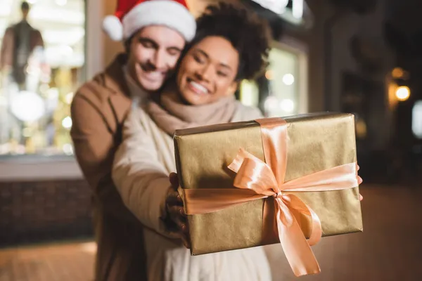 Mujer afroamericana borrosa y feliz sosteniendo caja de regalo cerca de novio en sombrero de santa - foto de stock