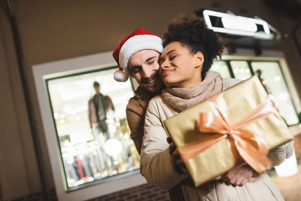 Heureuse femme afro-américaine tenant une boîte-cadeau floue près du petit ami dans le chapeau de Père Noël — Photo de stock