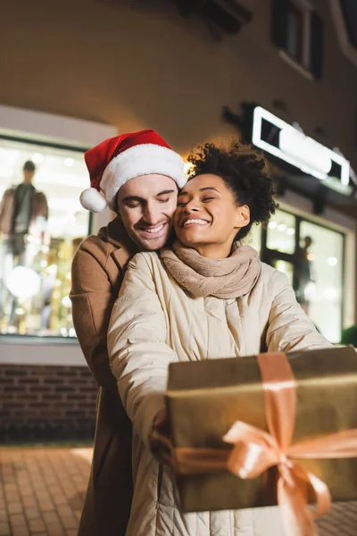 Felice donna africana americana in possesso di scatola regalo avvolto sfocato vicino fidanzato in cappello di Santa — Foto stock