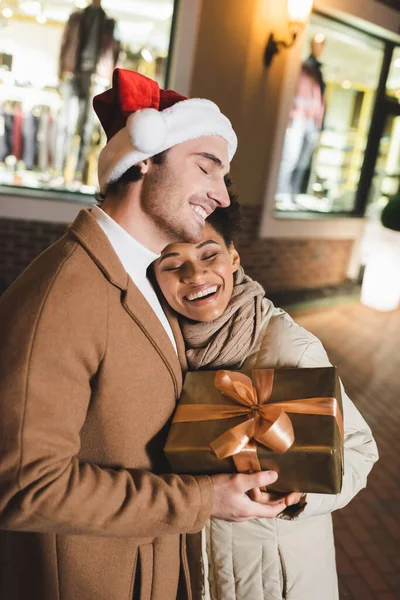 Mujer afroamericana feliz y hombre complacido en sombrero de santa celebración envuelto caja de regalo en la noche - foto de stock