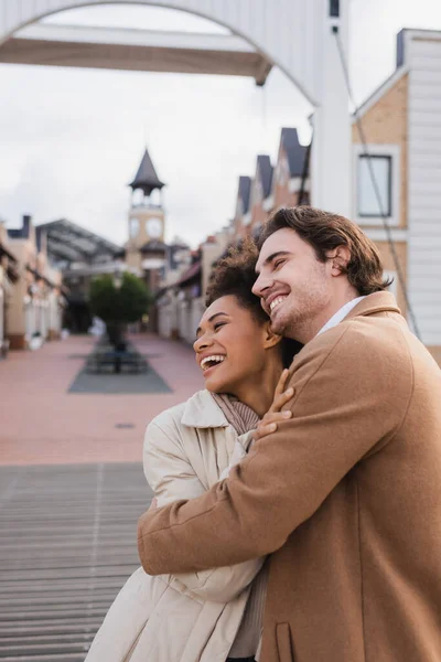 Joyful multiethnic couple in coats hugging near shopping center — Stock Photo