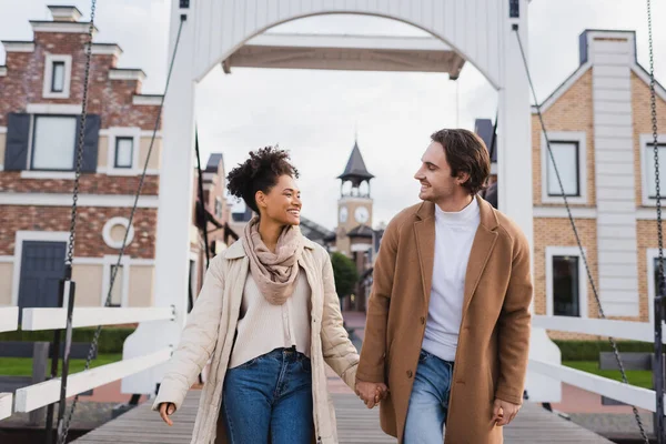 Smiling multiethnic couple in coats holding hands near shopping center — Stock Photo