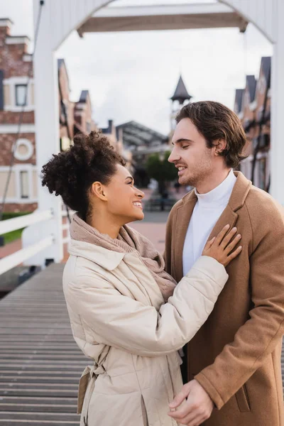 Smiling multiethnic couple in coats embracing near shopping center — Stock Photo