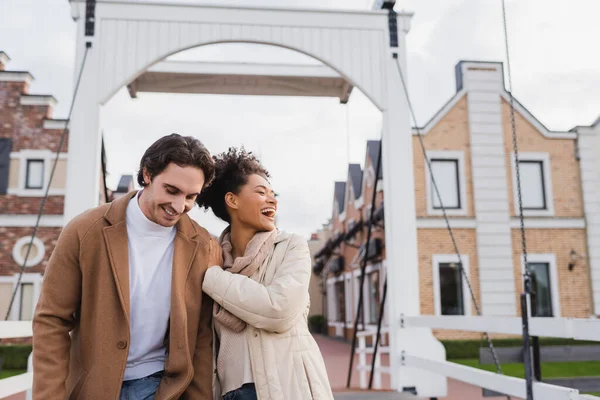 Pleased multiethnic couple in coats laughing near shopping center — Stock Photo