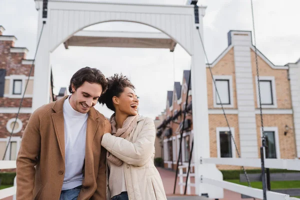 Heureux couple multiethnique en manteaux riant près du centre commercial — Photo de stock