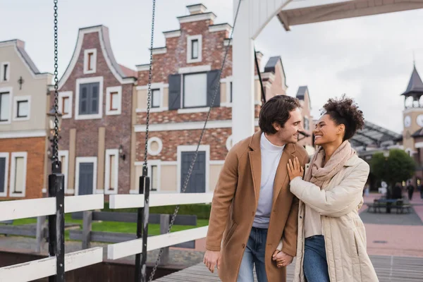 Joyful multiethnic couple in coats holding hands and smiling near shopping center — Stock Photo