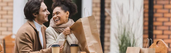 Happy african american woman holding shopping bag and laughing near boyfriend with smartphone, banner — Stock Photo