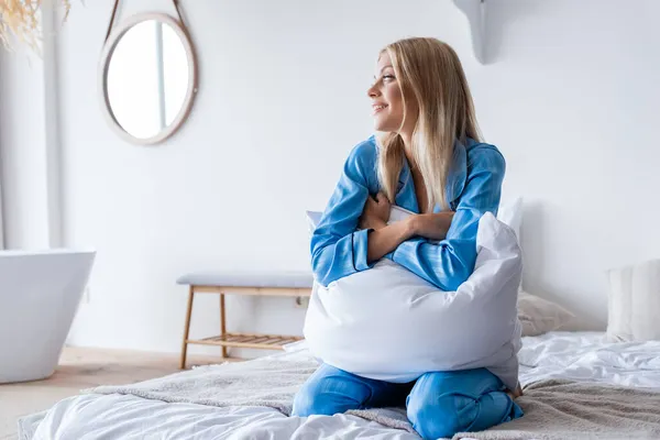 Alegre joven mujer sosteniendo almohada mientras mira hacia otro lado en el dormitorio - foto de stock