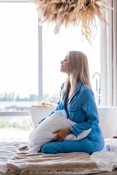 Vue latérale de la jeune femme joyeuse tenant oreiller tout en étant assis sur le lit — Photo de stock