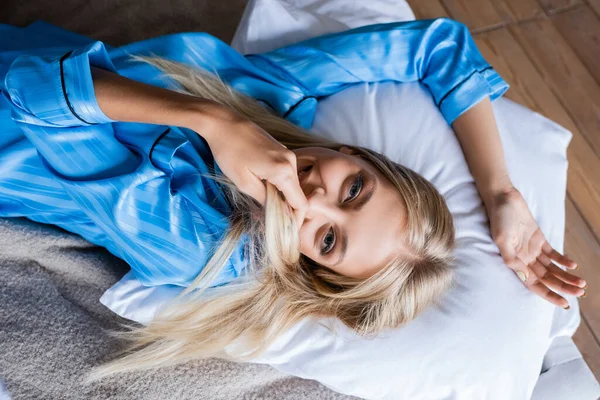 Вид сверху счастливой блондинки, закрывающей рот волосами, лежа на подушке в спальне — стоковое фото