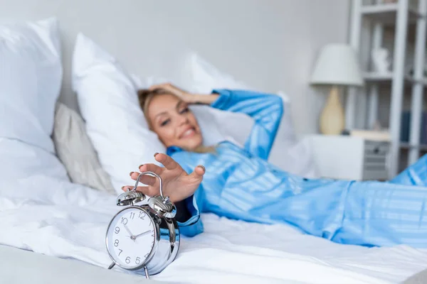 Mujer feliz y borrosa que alcanza el reloj despertador borroso en el dormitorio - foto de stock