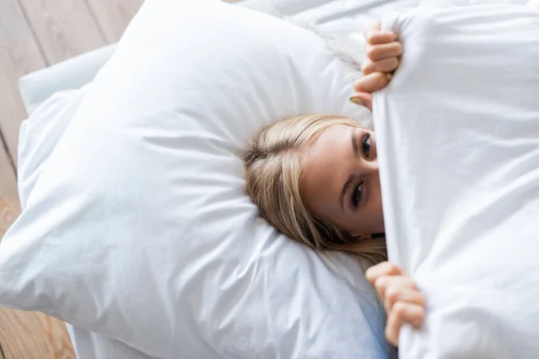 Top view of young woman covering face with blanket while lying on bed — Stock Photo