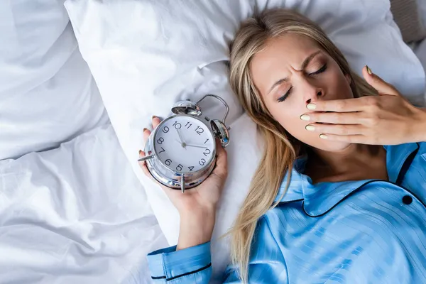 Draufsicht der Frau, die Mund und Gähnen bedeckt, während sie mit Wecker im Bett liegt — Stockfoto